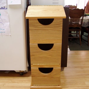 May include: A three-drawer wooden storage unit with a light wood finish. The drawers have a rounded cutout on the front. The unit is standing on a wood floor.