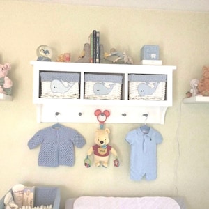 May include: White shelf unit with three baskets featuring a whale design. The shelf has hooks for hanging items. There are two blue knitted baby outfits hanging on the hooks. A Winnie the Pooh stuffed animal is hanging from a hook. The shelf is mounted on a light yellow wall.