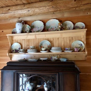 May include: A wooden shelf with a scalloped edge, holding a collection of vintage porcelain plates and teacups. The plates feature floral and fruit designs in shades of pink, purple, and green. The shelf is mounted on a wooden wall.