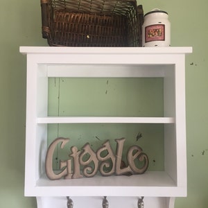 May include: White wooden shelf with two shelves and hooks on the bottom. A wicker basket and a white jar with a floral design are on the top shelf. The word "Giggle" is on the bottom shelf.