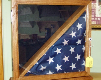 May include: A wooden shadow box with a glass front, displaying a folded American flag in a triangular shape. The box has a dark wood stain and is sitting on a light wood surface.