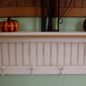 Puede incluir: Estante de madera blanca con un respaldo de tablilla decorativa y cuatro ganchos. El estante tiene un acabado pintado en blanco y está decorado con dos pequeñas calabazas.