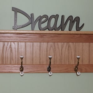 Może przedstawiać: Drewniany wieszak na ubrania z półką i pięcioma haczykami. Półka ma ozdobną listwę, a haczyki mają białe porcelanowe gałki. Na półce widnieje napis "Dream".