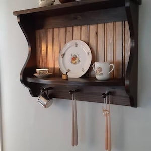 May include: A dark brown wooden wall shelf with a curved top and a lower shelf with hooks. The top shelf has a white and gold patterned plate, a small white teacup, and a small gold object. The lower shelf has a silver cup and two pairs of long, thin, light pink candles.