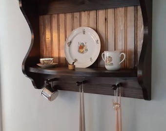 Puede incluir: Una estantería de pared de madera marrón oscuro con la parte superior curvada y una repisa inferior con ganchos. La repisa superior tiene un plato con un patrón blanco y dorado, una pequeña taza de té blanca y un pequeño objeto dorado. La repisa inferior tiene una taza de plata y dos pares de velas largas, delgadas y de color rosa claro.