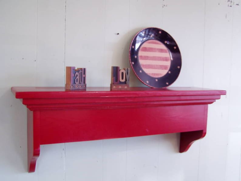 Puede incluir: Una estanter&iacute;a de madera roja con un borde decorativo. La estanter&iacute;a est&aacute; montada en una pared blanca y tiene un plato rayado azul y blanco con estrellas, y dos bloques de madera que deletrean "faith" y "joy" en la parte superior.
