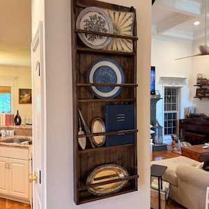 May include: A tall, dark brown wooden wall-mounted display shelf with vertical beadboard backing. It features five horizontal rails holding two decorative plates, a dark blue book, a silver-toned serving utensil, and two oval metal trays.