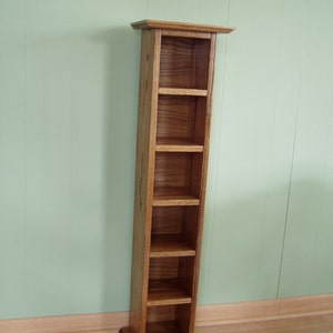 May include: A wooden DVD or CD storage tower with five shelves. The tower is made of light brown wood with a natural wood grain finish.