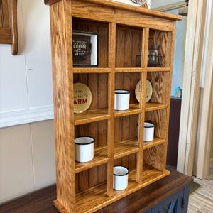 May include: A wooden wall shelf with multiple compartments, stained in a warm brown hue. The shelf contains small white enamel mugs with black rims, decorative plates, and framed artwork with the text "Coffee and Jesus".
