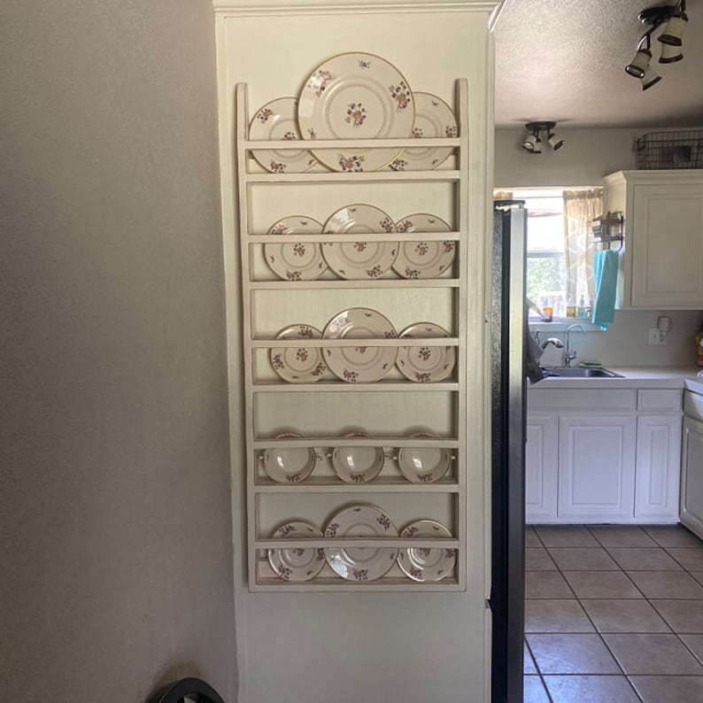 May include: A white wooden plate rack with 8 shelves holding white plates with floral designs. The rack is mounted on a white wall in a kitchen.
