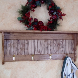 May include: A wooden coat rack with a shelf and five hooks. The rack is stained a dark brown and has a rustic look. A green and white towel is hanging on one of the hooks. A red and green Christmas wreath with red apples is hanging on the wall above the rack.