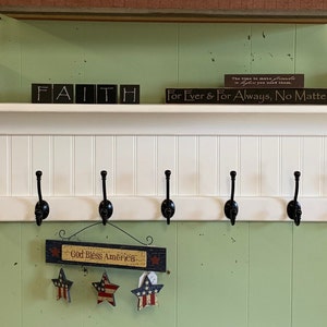 Op de afbeelding: Wit houten plank met een wit schuttingsontwerp en acht zwarte metalen haken. De plank heeft een decoratieve rand aan de voor- en achterkant. De plank is gemonteerd aan een groene muur. Er staan ​​verschillende kleine decoratieve dozen en borden op de plank, waaronder een met de tekst "Geloof" en een andere met de tekst "Voor altijd en eeuwig, wat er ook gebeurt".
