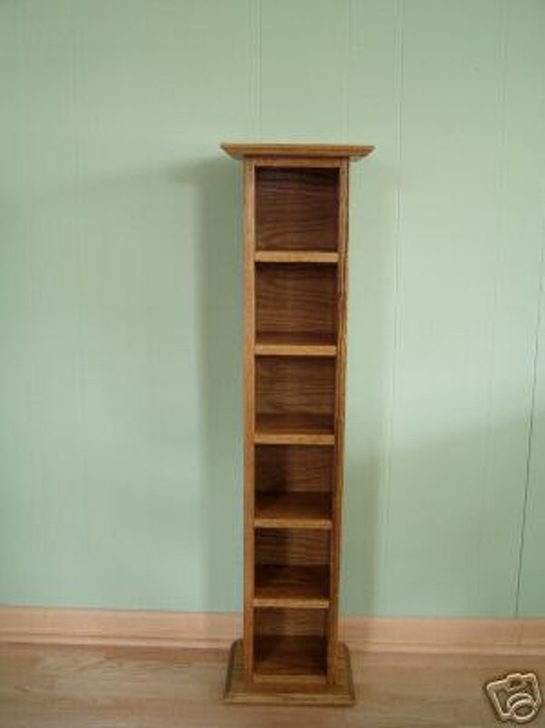 May include: A freestanding wooden DVD storage tower with six shelves. The tower is made of light brown wood with a natural wood finish.