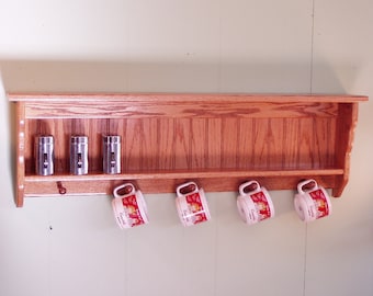 May include: A wooden wall shelf with a decorative, paneled front. The shelf has three spice jars and four mugs hanging from hooks on the bottom.