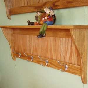 May include: A wooden coat rack with a shelf and five hooks. The rack is made of light brown wood and has a slightly distressed finish. The shelf is about 12 inches deep and the hooks are about 4 inches long. The rack is mounted on a light green wall.