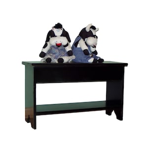 May include: Black wooden bench with a shelf, featuring two plush cow toys sitting on top. The cow toys are black and white with a red mouth and blue accents.