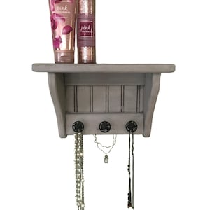 May include: A gray wooden shelf with three black hooks, holding necklaces and two bottles of body care products. The bottles are labeled "pink cashmere".