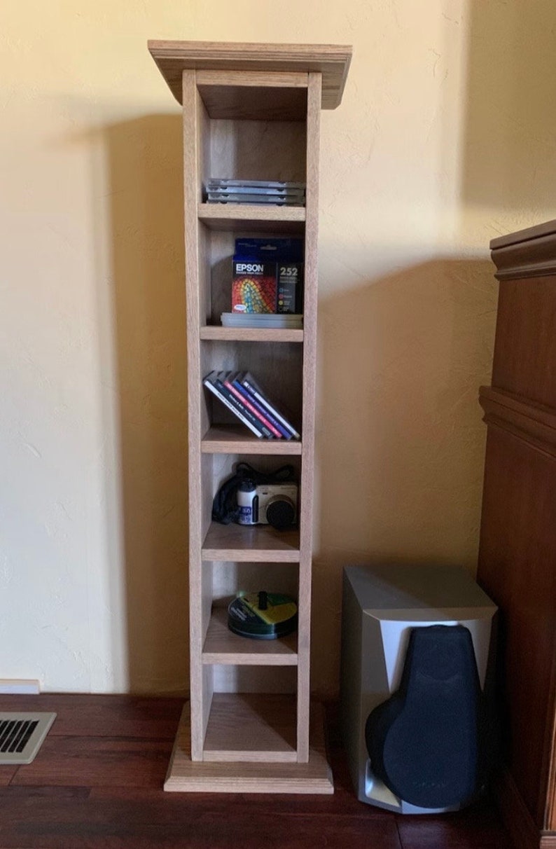 May include: A wooden CD tower with six shelves, filled with CDs, ink cartridges, and a camera. The tower is standing on a wooden floor next to a black speaker.