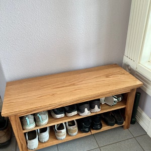 May include: A wooden shoe rack with two shelves. The shoe rack is made of light brown wood and has a natural finish. The shoe rack is filled with shoes, including sneakers, sandals, and dress shoes.