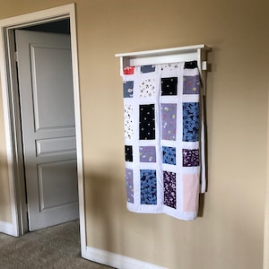 May include: A white quilt rack with a white shelf and a patchwork quilt hanging from it. The quilt is made of various fabrics in shades of blue, white, black, and pink. The quilt is hung on the rack with a white fabric backing.