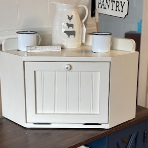May include: A white farmhouse-style bread box with a hinged door and a small knob. A white pitcher with black farm animal silhouettes sits on top, along with two white enamel mugs. A small sign that says "Faith" is also on top.