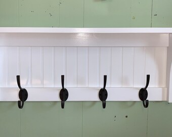 May include: White wooden shelf with four black metal hooks. The shelf has a decorative, slightly curved, top edge and a beaded board back.