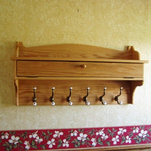 May include: A wooden wall shelf with a single drawer and six hooks. The shelf is stained a light brown color and has a slightly curved top edge. The hooks are made of metal and have white porcelain knobs.