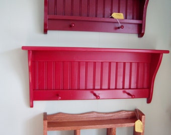 May include: Two red wooden shelves with a scalloped edge and three hooks on the bottom shelf. The shelves are mounted on a white wall.