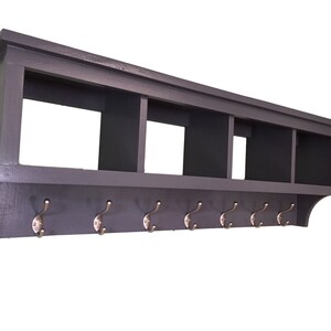May include: A dark gray wooden wall shelf with a built-in top shelf and seven brass-colored hooks. The shelf has three open compartments and a curved bottom edge.