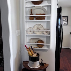 May include: A white wall-mounted shelf unit displaying vintage kitchenware. Items include wooden bowls, plates with floral designs, rolling pins, and a ceramic crock. The shelf is positioned in a corner, with a dark wood floor visible.
