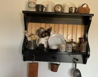 Puede incluir: Una estantería de pared negra con un panel trasero de madera, que muestra varios artículos de cocina vintage. Incluye un reloj, tazas, platos, tarros y una pequeña cesta de mimbre. Una tabla de cortar de madera y un cucharón de metal cuelgan debajo.
