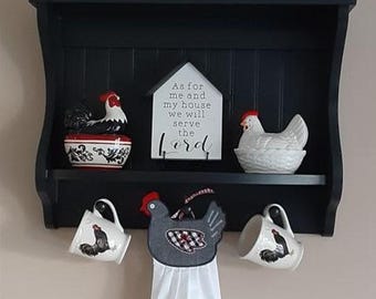 May include: A black wooden shelf with two shelves. The top shelf has a white sign with the text "As for me and my house we will serve the Lord". The bottom shelf has two white mugs with black rooster designs, a white ceramic rooster, and a gray and black chicken dish towel.