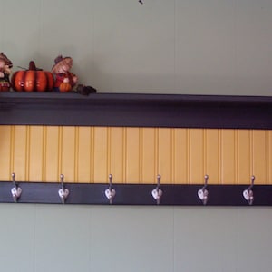 Puede incluir: Un estante de pared negro y amarillo con seis ganchos plateados. El estante tiene una parte superior e inferior negras con un centro amarillo. Dos figuras y una calabaza se encuentran en la parte superior.