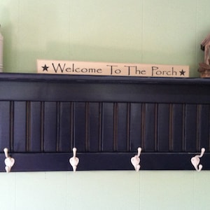 Puede incluir: Una estantería de madera azul oscuro con un letrero blanco que dice "Welcome To The Porch" y cuatro ganchos blancos. Un pequeño frasco blanco y una casita de pájaros están en la estantería.