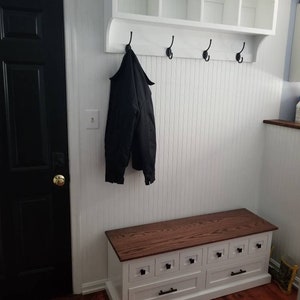 Puede incluir: Un banco de entrada blanco con una tapa de madera marr&oacute;n y seis cajones. Encima del banco hay un estante blanco con cuatro cub&iacute;culos y cuatro ganchos negros. Un abrigo negro cuelga de uno de los ganchos. La puerta es negra.