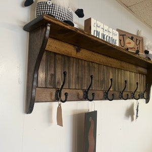 May include: A wooden wall-mounted shelf with a brown stain and a decorative trim. The shelf has six black metal hooks and a small shelf on top. The shelf is decorated with a snowman figurine, a small sign that says "Joy", and a sign that says "I find my purpose better, I'm negative".