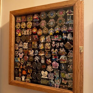 May include: A wooden shadow box with a glass front filled with a collection of colorful Disney character pins. The pins are arranged in rows and columns on a black background.