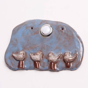 May include: A ceramic doorbell with a blue and brown glaze. The doorbell features a white round button and four small white birds with brown legs.