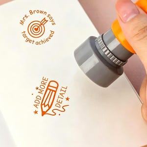 Könnte beinhalten: Ein grauer und orangefarbener Selbstfärbestempel, der orange Tinte auf weißes Papier druckt. Der Stempel erzeugt zwei Motive: ein Zielscheibensymbol mit dem Text "Mrs. Brown says target achieved" und ein Bleistiftsymbol mit "ADD MORE DETAIL".