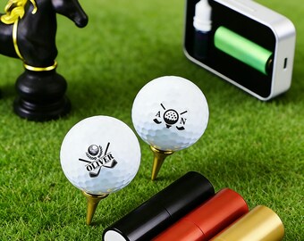 Könnte beinhalten: Zwei weiße Golfbälle mit schwarzen Logos und dem Namen "OLIVER" auf goldenen Tees, platziert auf grünem Gras. Eine schwarze und goldene Schachfigur und ein silbernes Gerät mit einer grünen Batterie befinden sich im Hintergrund. Drei farbige Tuben sind im Vordergrund.