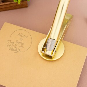May include: Gold-toned metal stamp with a round design featuring the text "Albert & Betty 02.12.2025" surrounded by a floral motif. The stamp is being used to create an impression on a brown card.