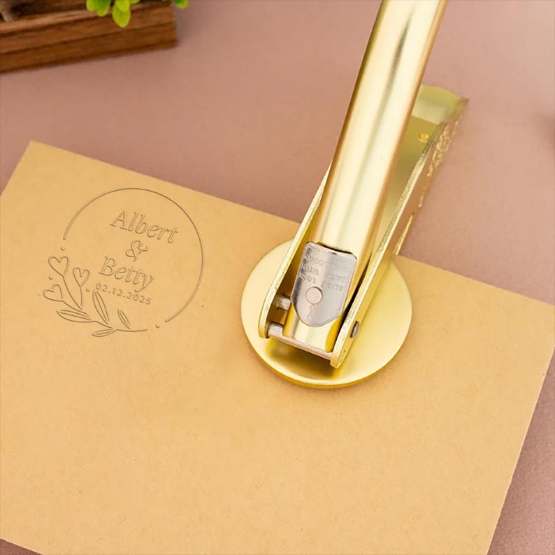 May include: Gold-toned metal stamp with a round design featuring the text "Albert & Betty 02.12.2025" surrounded by a floral motif. The stamp is being used to create an impression on a brown card.