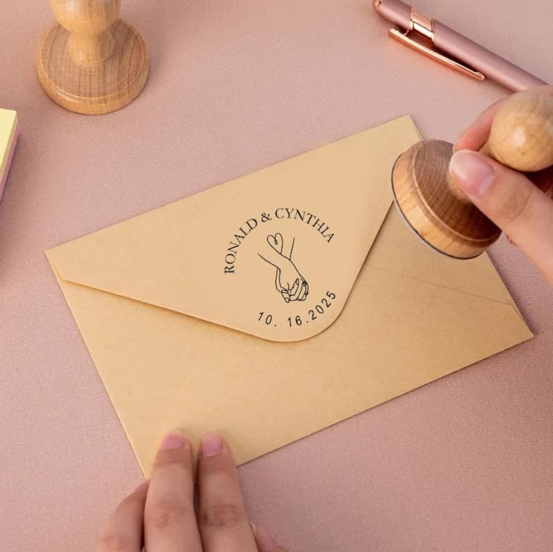 May include: A brown envelope with a custom rubber stamp. The stamp features a line drawing of two hands holding each other with a heart in the middle. The text on the stamp reads "Ronald & Cynthia 10.16.2025".