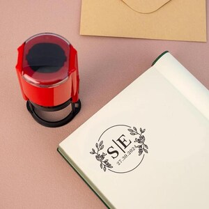 May include: A red self-inking stamp with a clear top and a black base. The stamp features a circular design with the letters "S|E" and the date "27.30.2024" inside a circle with floral accents.