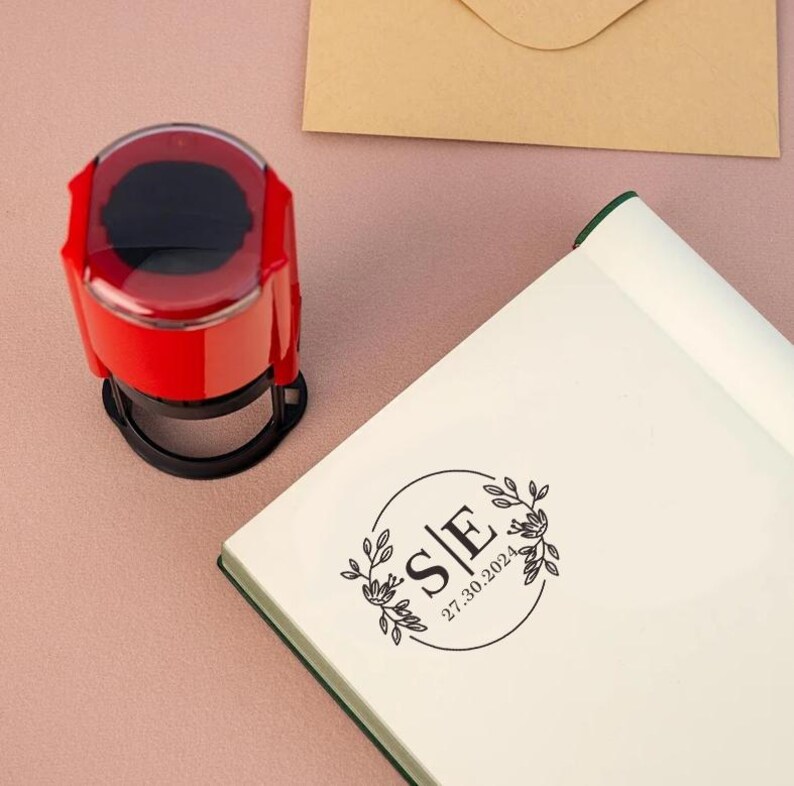 May include: A red self-inking stamp with a clear top and a black base. The stamp features a circular design with the letters "S|E" and the date "27.30.2024" inside a circle with floral accents.