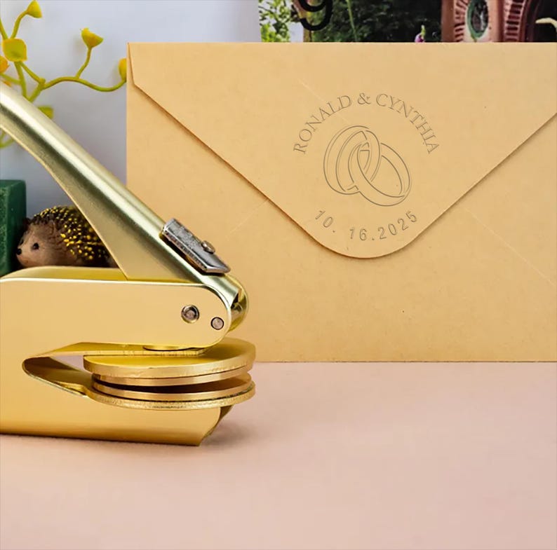 May include: A gold-colored self-inking stamp with a round design featuring two interlocking rings and the text "RONALD & CYNTHIA 10.16.2025".