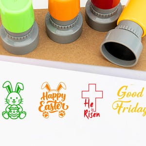 Op de afbeelding: Een set kleurrijke paasstempels met groene, oranje, rode en gele handvatten. De stempels bevatten een afbeelding van een konijn, de tekst "Happy Easter", een kruis met "He is Risen" en "Good Friday."