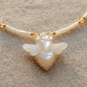 May include: A necklace featuring a carved mother-of-pearl bee pendant suspended from a strand of small, cylindrical white beads interspersed with tiny, round, translucent golden-yellow beads and small metallic gold-toned spacer beads.