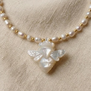 May include: A necklace featuring a central pendant carved from iridescent white mother-of-pearl in the shape of a bee with detailed wings. The chain consists of small, oval-shaped white freshwater pearls interspersed with tiny, round gold-tone beads.