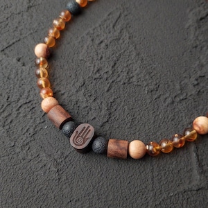 Puede incluir: Collar de cuentas con cuentas de color ámbar, de madera y de piedra volcánica oscura. Incluye un dije de madera con un símbolo de mano. Estilo natural y terroso.